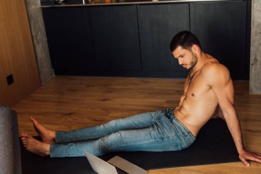 Üstsüz bir adam yoga minderinde oturup laptopta yoga eğitimi alıyor. 