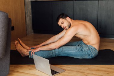 Üstsüz adam yoga minderinde pratik yapıyor ve laptopta yoga eğitimi alıyor. 