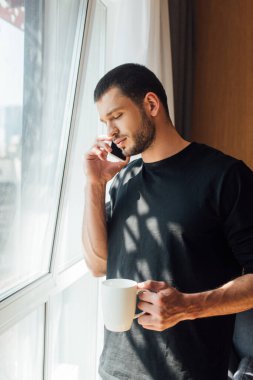 Evde akıllı telefonla konuşurken elinde bardak tutan sakallı adam.