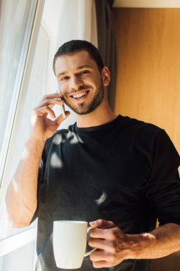 Evde elinde bardak tutan ve akıllı telefondan konuşan mutlu bir adam.