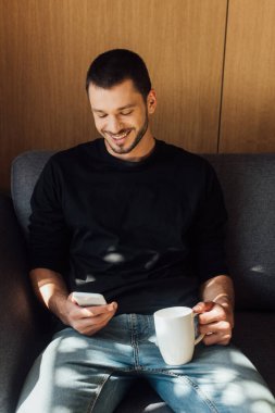 Güneş ışığı, sakallı adamın gülümserken akıllı telefon kullanması ve oturma odasında bardak tutması. 