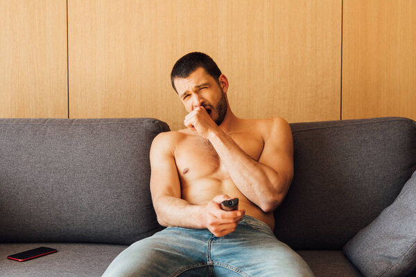 tired shirtless man in jeans yawning and holding remote controller 