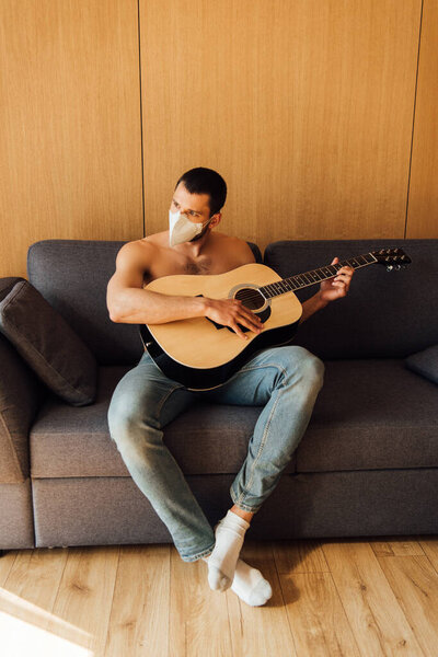 shirtless man in medical mask playing acoustic guitar in living room 