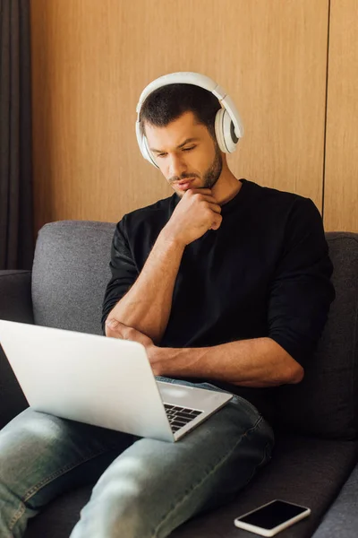 pensive freelancer in wireless headphones looking at laptop near smartphone with blank screen on sofa