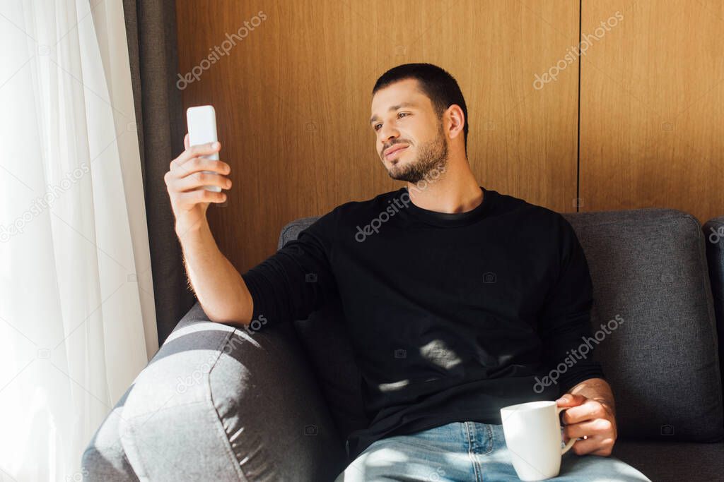 sunlight on handsome man smiling while using smartphone and holding cup in living room 