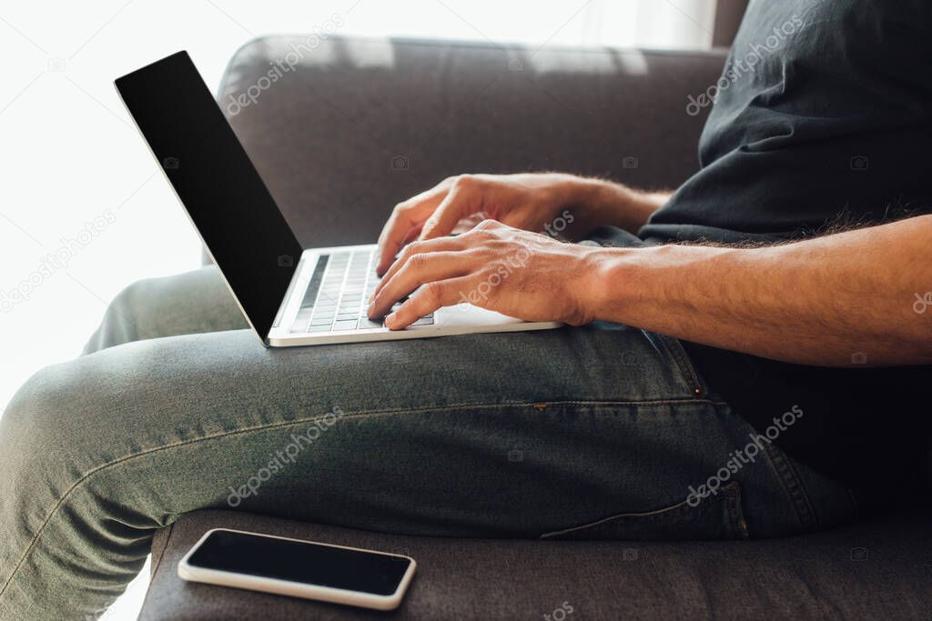 cropped view of freelancer typing on laptop keyboard near smartphone with blank screen on sofa 