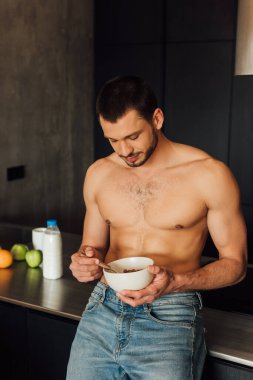 Tişörtsüz adam mutfakta mısır gevreği ve kaşıkla kase tutuyor.  