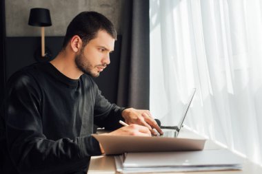 Laptop kullanan ve evde klasörün yanında kalem tutan sakallı serbest çalışanın seçici odak noktası 
