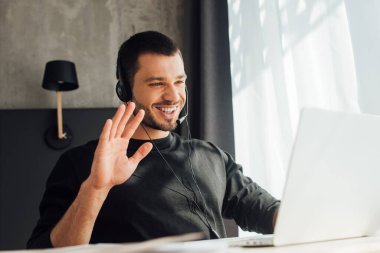 selective focus of happy operator in headset waving hand while having video call at home clipart