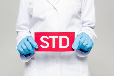 cropped view of doctor in blue latex gloves and white coat holding std lettering isolated on grey clipart