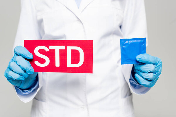 cropped view of doctor in white coat and latex gloves holding paper with std lettering and condom isolated on grey 