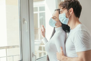 Tıbbi maskeli hasta bir çift, tecrit sırasında pencereden dışarı bakıyor.