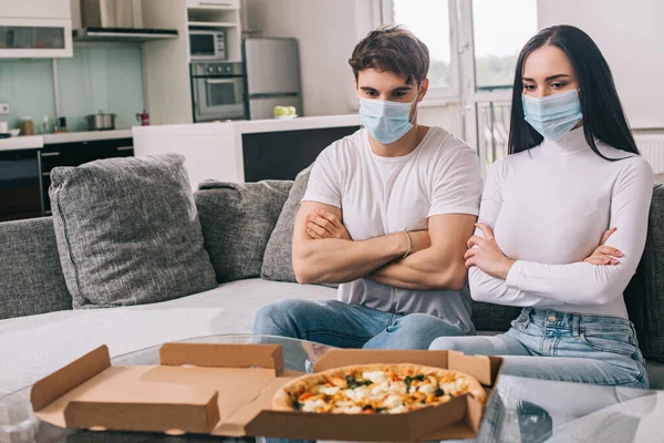 自己分離中にピザを家で見て腕を交差させた医療用マスクの悲しい病気のカップル — ストック写真