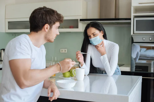 man and ill woman in medical mask looking at thermometer and having breakfast during self isolation