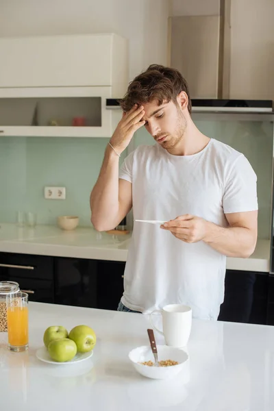 worried ill man with fever looking at thermometer on kitchen during breakfast on self isolation