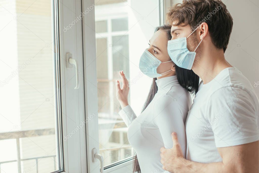 ill couple in medical masks looking through window during self isolation