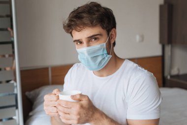 Tıbbi maskeli genç adam kendini izole ederken sıcak içecekle bardağı yatakta tutuyor.