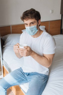 Tıbbi maskeli yorgun bir adam kendini izole ederken sıcak içecekle bardağı yatakta tutuyor.