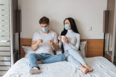 Tıbbi maskeli endişeli çift, kendilerini izole ettikleri sırada yatak odasında bardak ve termometre tutuyorlar.