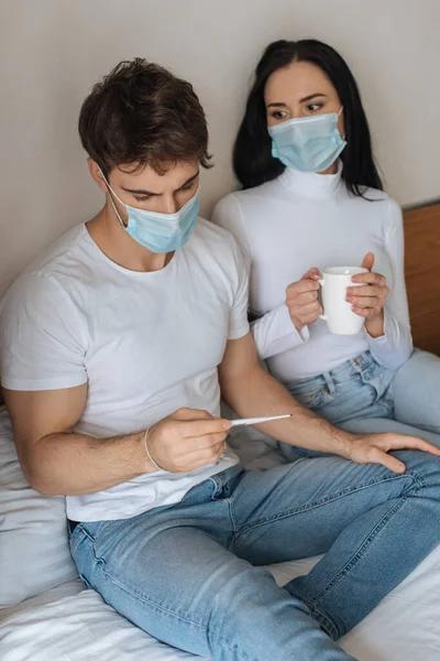 戴医疗面罩的生病夫妇在自我隔离时将杯子和温度计放在床上 — 图库照片