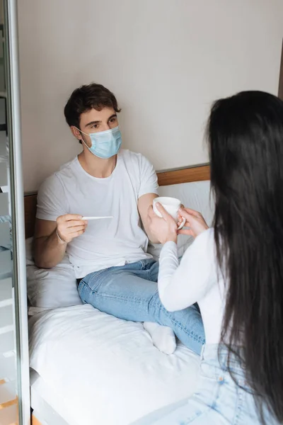 女性は自己分離の間にベッドの温度計で医療用マスクの男に熱い飲み物を与える — ストック写真