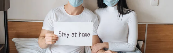 cropped view of ill couple in medical masks holding Stay at home sign at home during self isolation, horizontal crop