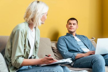 Kanepede dizüstü bilgisayar kullanan ve sarı duvarın yanındaki not defterine yazı yazan serbest çalışan genç bir çift.