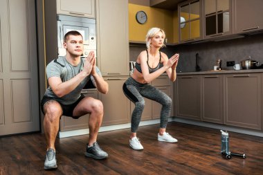 young couple doing squats together at home during quarantine clipart