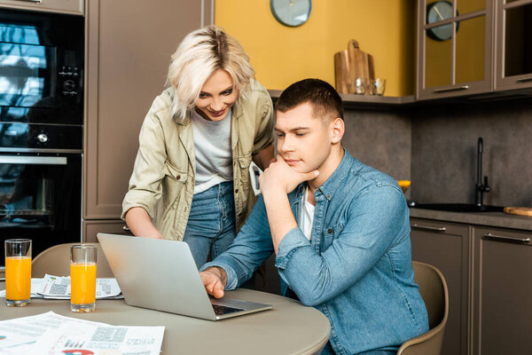 улыбающаяся пара, работающая с бумагами и ноутбуком возле апельсинового сока в газах на кухне дома
