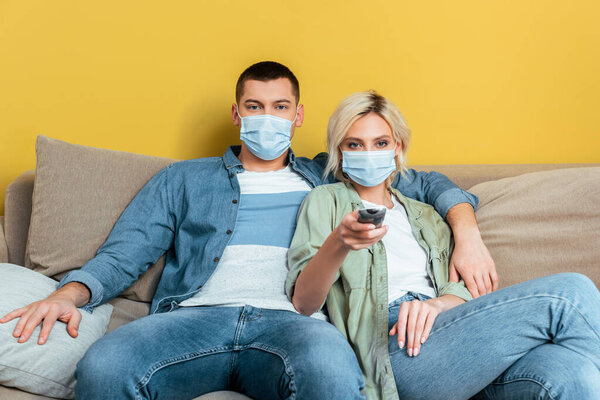 young couple in medical masks watching tv on sofa near yellow wall