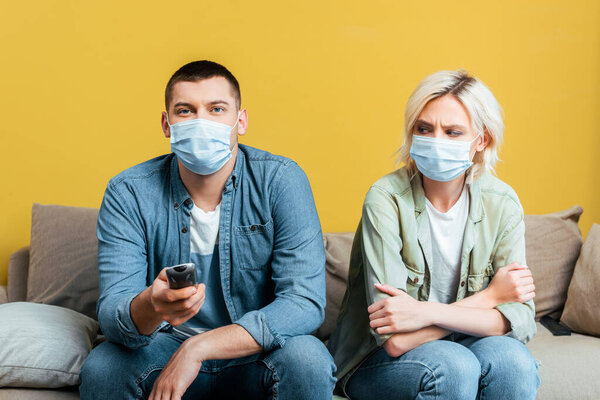 young man in medical mask watching tv near offended girlfriend on sofa near yellow wall