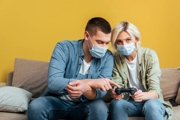 KYIV, UKRAINE - APRIL 22, 2020: young man in medical mask teaching girlfriend playing video games on sofa near yellow wall