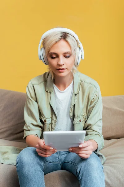 Młoda Blondynka Słuchawkach Tabletem Cyfrowym Kanapie Pobliżu Żółtej Ściany — Zdjęcie stockowe