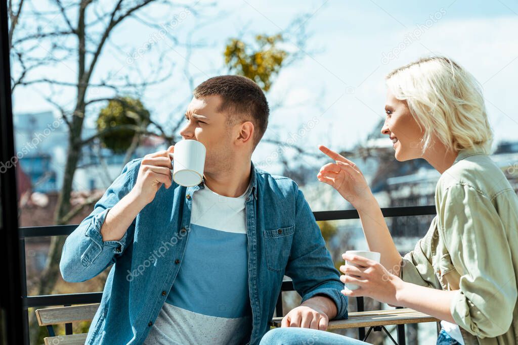 smiling couple talking and drinking tea on terrace while woman pointing with finger away