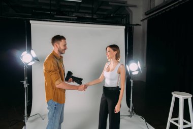 Side view of photographer holding digital camera while shaking hands with smiling model in photo studio  clipart