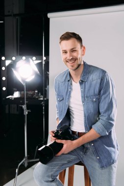 Gülümseyen fotoğrafçı fotoğraf stüdyosunda projektörün yanında dijital kamera tutuyor. 