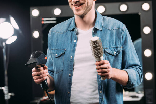 Cropped view of smiling makeup artist holding hairbrush and hair dryer in photo studio 