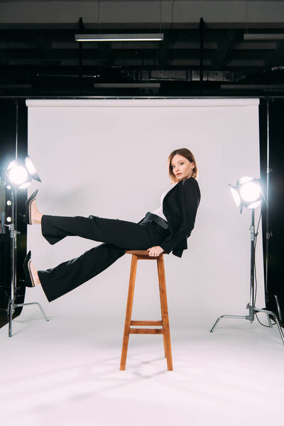 Side view of stylish model looking at camera while sitting on chair in photo studio 
