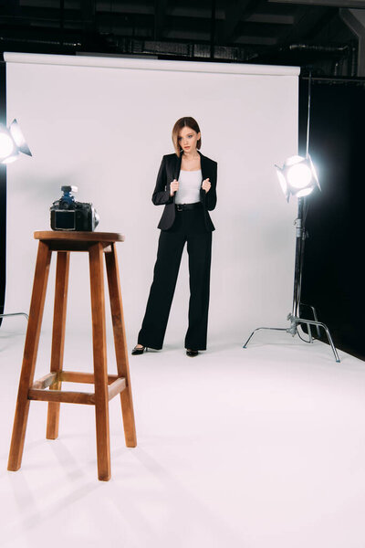 Attractive elegant model posing at digital camera on wooden chair in photo studio 
