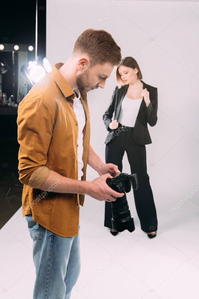 Selective focus of photographer looking at display of digital camera near elegant model and floodlight in photo studio 