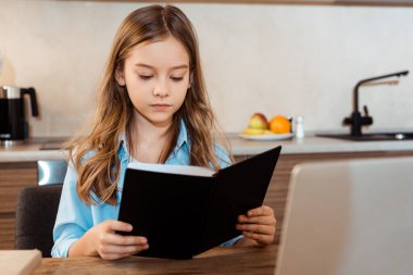 Evde e-öğrenim görürken dizüstü bilgisayarın yanında kitap okuyan sevimli bir çocuğun seçici odağı