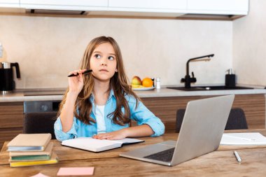 Evde e-öğrenim görürken dizüstü bilgisayarın yanında kalem tutan düşünceli çocuğun seçici odağı