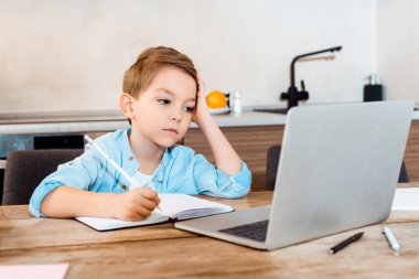 Dizüstü bilgisayara bakarken ve evde e-öğrenirken not defterine yazmaktan sıkılan çocuğun seçici odağı