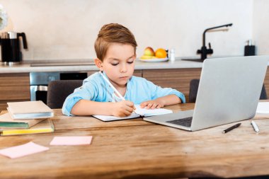 selective focus of boy writing in notebook near laptop while e-learning at home clipart