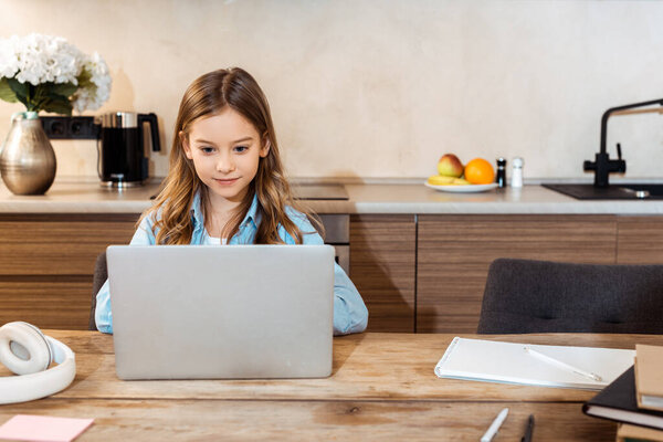cute kid online studying while looking at laptop near headphones and notebook at home