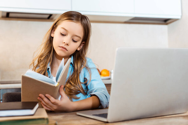 selective focus of cute kid holding book near laptop while online studying at home