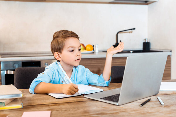 selective focus of boy writing in notebook and looking at laptop while e-learning at home