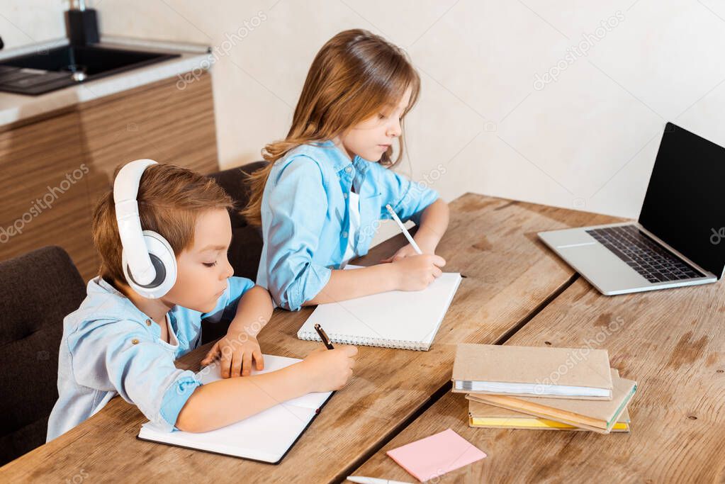 siblings writing in notebooks while e-learning near laptop with blank screen