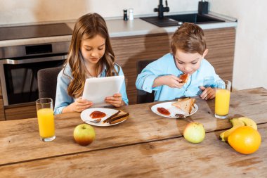 Dijital tablet kullanan kız kardeşinin yanında reçelli tost yiyen çocuk. 