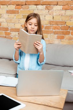 Laptop ve dijital tabletin yanında kitap okuyan sevimli çocuğun seçici odak noktası